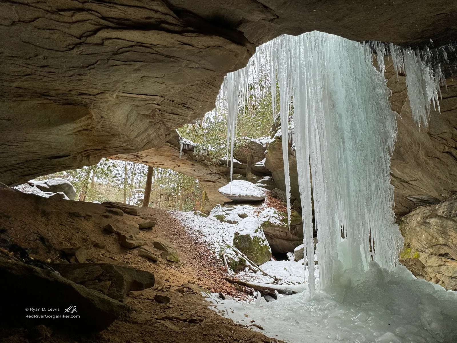 Large layered ice column beneath the sandstone at Red-byrd Arch, surrounded by snow-covered rocks and winter forest.