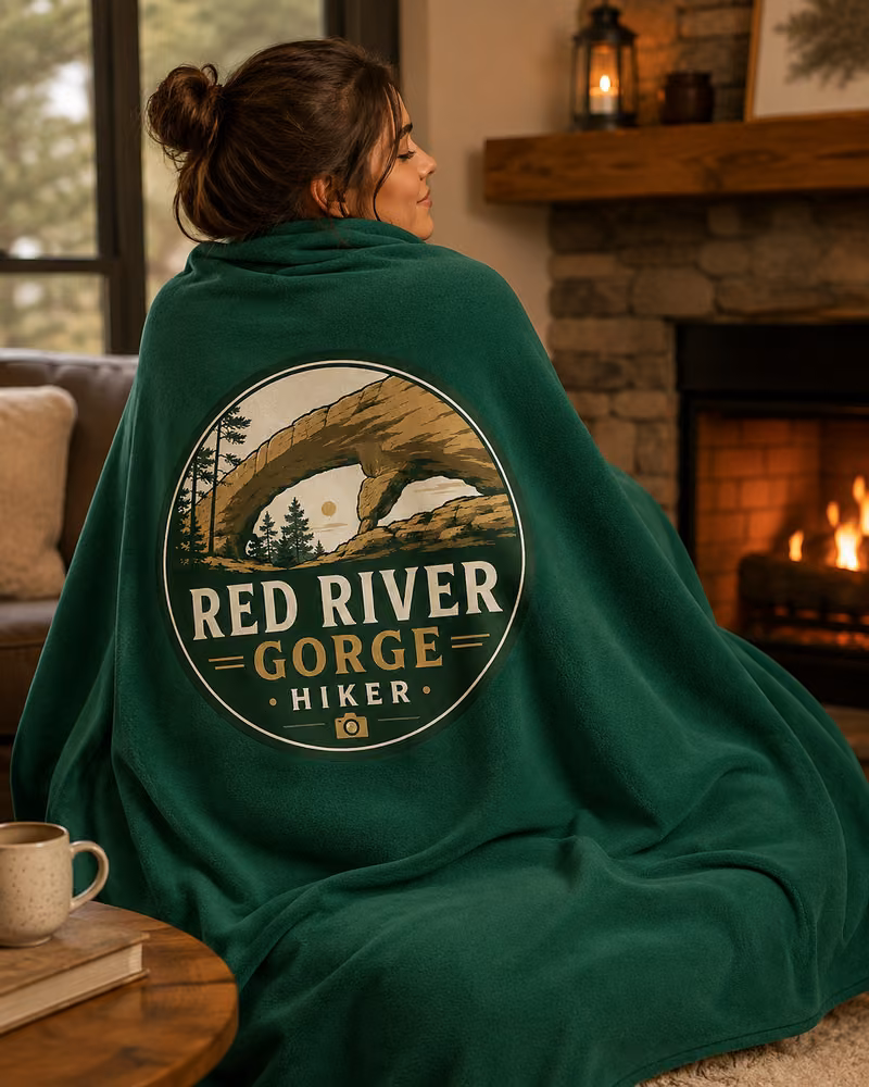 Person wrapped in a green Red River Gorge Hiker logo fleece blanket beside a fireplace.